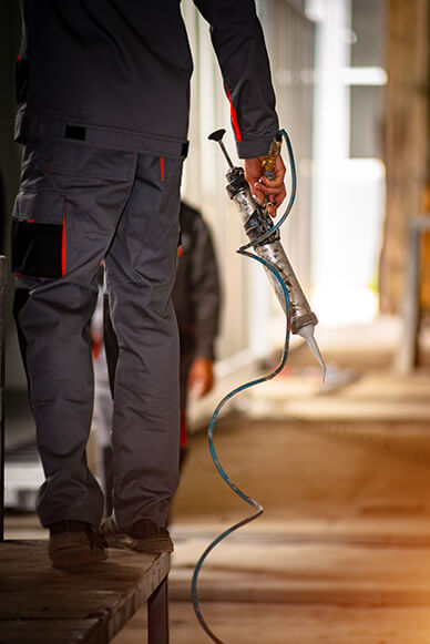 Worker in grey protective clothing holding a caulking gun with a connected hose, standing on a wooden platform in an industrial setting.