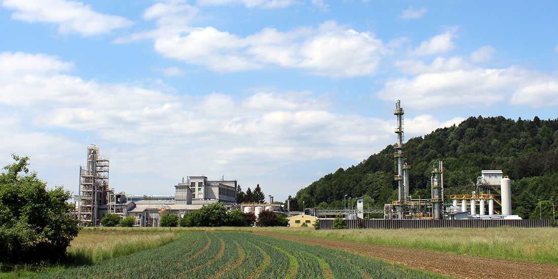 Industrial facility with tall structures and multiple buildings, set in a rural landscape with crop fields in the foreground and forested hills in the background.