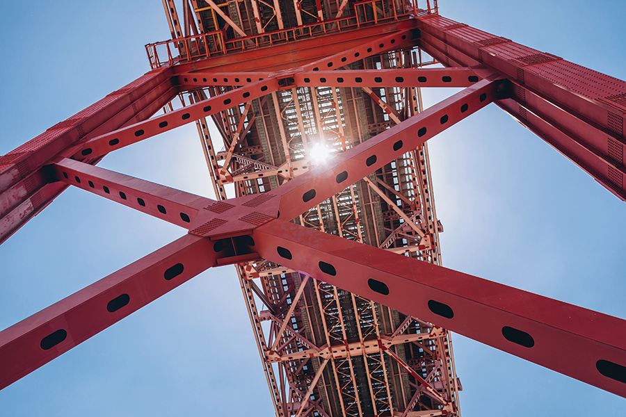 Close-up view of a red steel bridge structure with sun shining through beams against a clear blue sky.