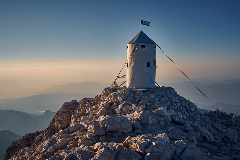 Cultural-heritage-preservation A small white cylindrical shelter with a flag stands atop a rocky mountain peak at sunrise, overlooking a vast mountain landscape. The structure is secured with cables and symbolizes cultural heritage preservation.