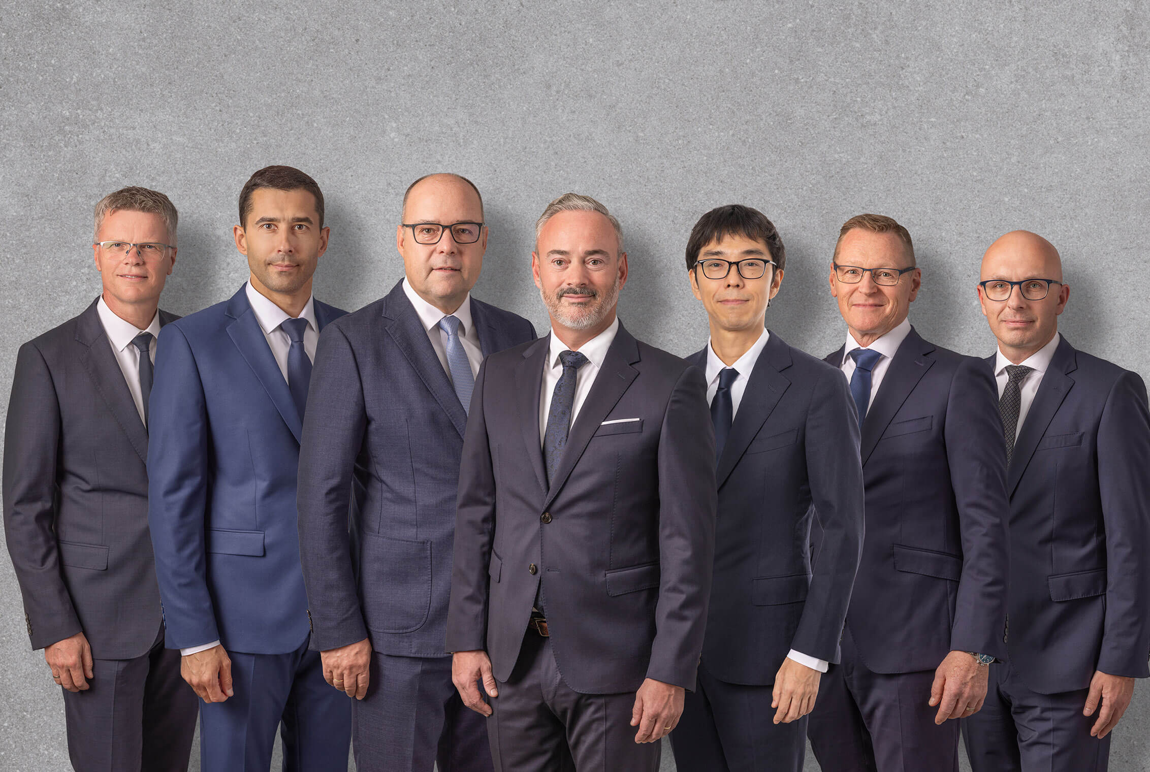 Group photo of eight men dressed in formal business suits, standing side by side in a row against a neutral gray background. They are positioned in a professional, upright manner, conveying a serious and corporate atmosphere. The coordinated attire and setting create a cohesive and executive impression.