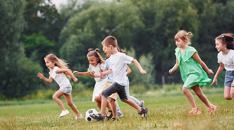 Kansai_Helios_Sustainability_Goals_02-e1723474327975 Group of young children playing football on a grassy field, smiling and running together, surrounded by trees in the background.