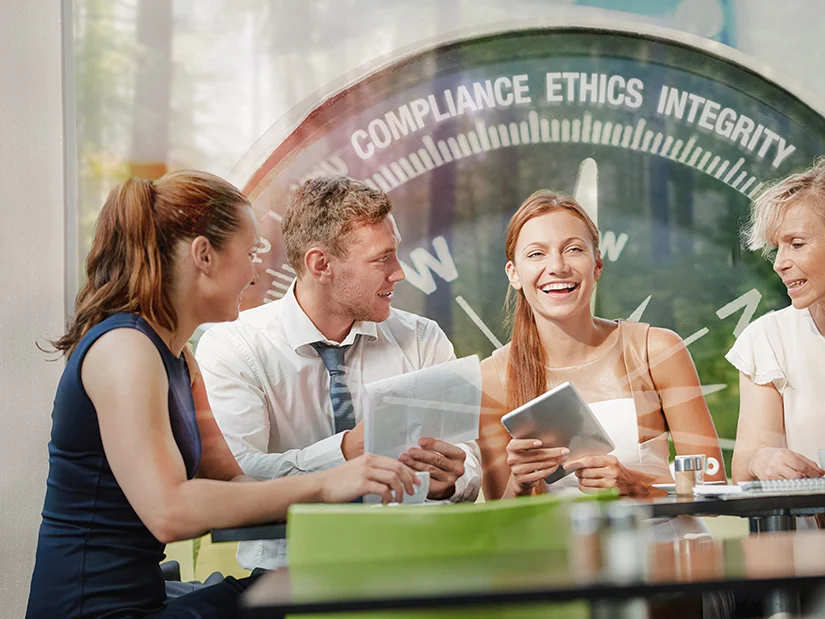 Group of professionals in a meeting, smiling and discussing documents, with a transparent overlay of a compass featuring the words ‘Compliance’, ‘Ethics’, and ‘Integrity’.