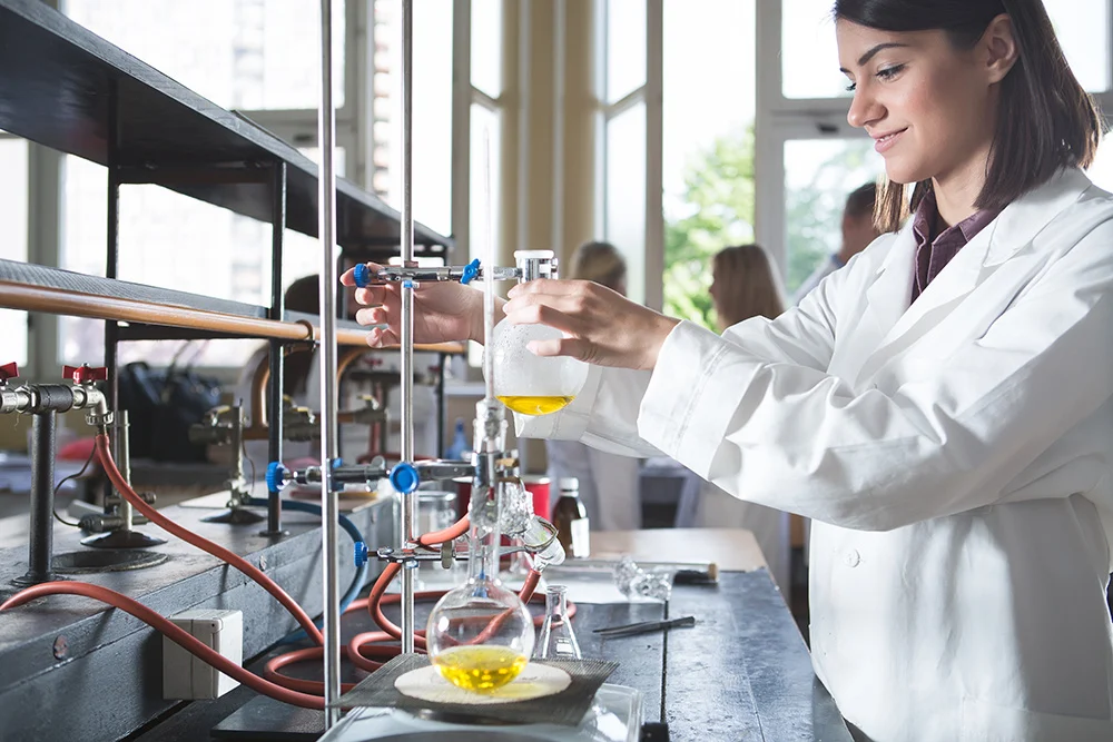 Lab_rd Young female scientist wearing a white lab coat conducting an experiment with laboratory glassware and yellow liquid in a bright, modern lab setting