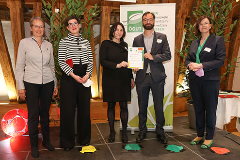 Five individuals stand on a stage, one holding a certificate, in front of a banner with the "ÖGUT" logo and greenery.