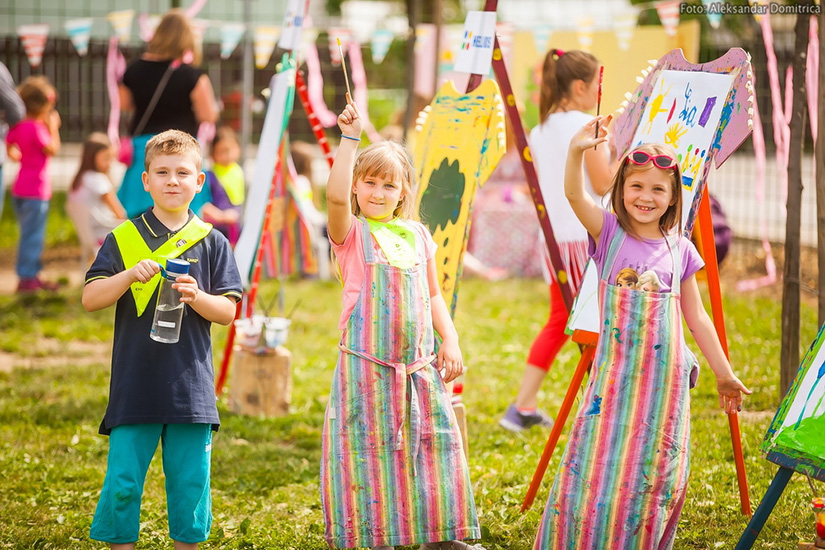 Rainbow-of-smiles Three smiling children in colorful aprons hold up paintbrushes during an outdoor painting activity. Behind them, easels display bright artwork as more children participate in the creative event on a sunny day