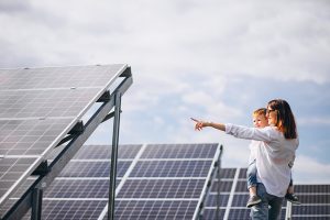 A person holding a child points toward rows of angled solar panels under a partly cloudy sky.