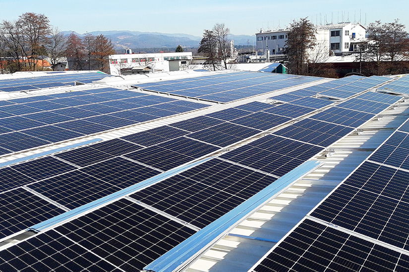 Solar-Power-Plant-02-825×550-Web-ENG The image shows a rooftop covered with solar panels. In the background, there are buildings, trees, and mountains under a clear sky.