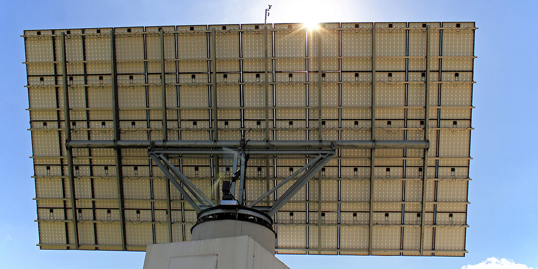 Underside view of a large solar panel array mounted on a metal framework. Panels are arranged in a grid pattern against a clear blue sky.