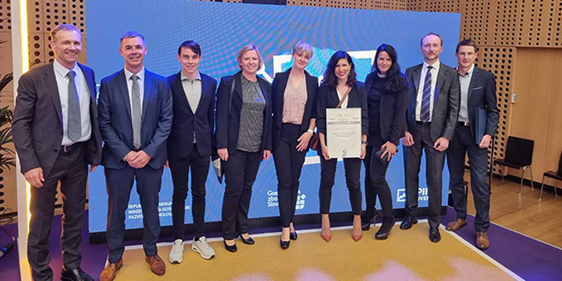 Nine people standing indoors in front of a screen. One person is holding a certificate, suggesting an award or recognition event.