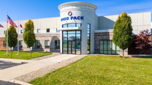 A modern building with typographic logo "OHIO PACK" on the front, featuring stone and panelled exterior. Three flags stand in front, with a glass entrance and landscaped surroundings.
