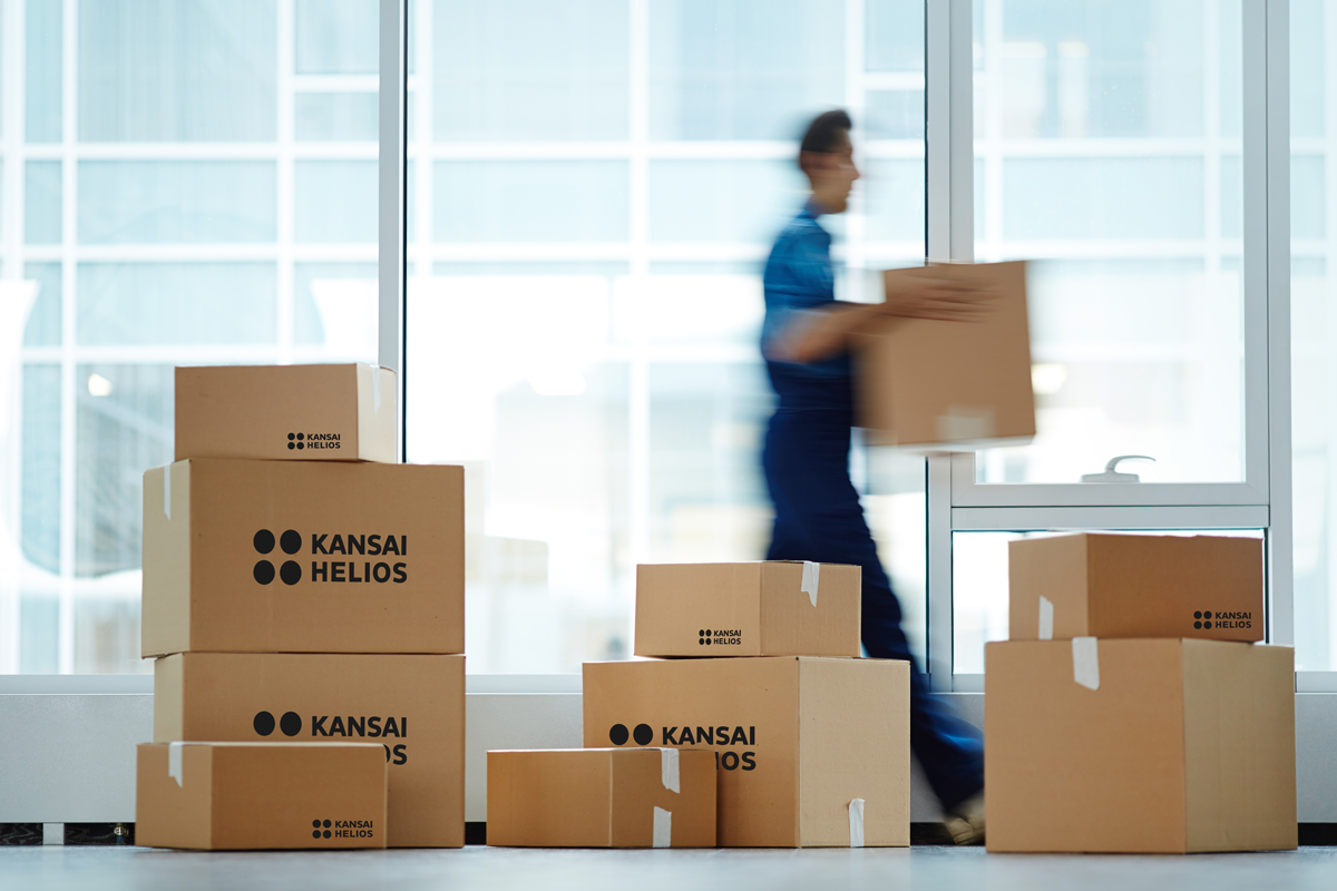 Moving situation with packed boxes branded with the KANSAI HELIOS logo and a moving company employee carrying away a box.