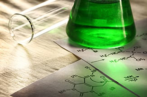 Close-up of a laboratory scene with a green liquid in a conical flask, a glass test tube, and chemical structure diagrams on paper under soft lighting.