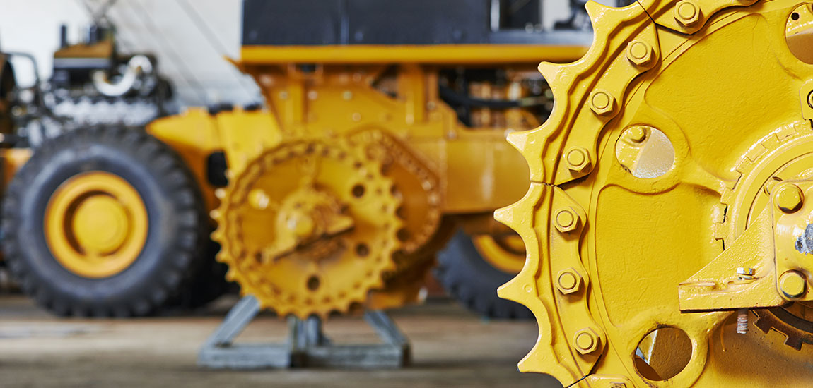 Close-up of a yellow sprocket and gear mechanism on a large industrial machine. Blurred background shows parts of similar machinery and large tyres.