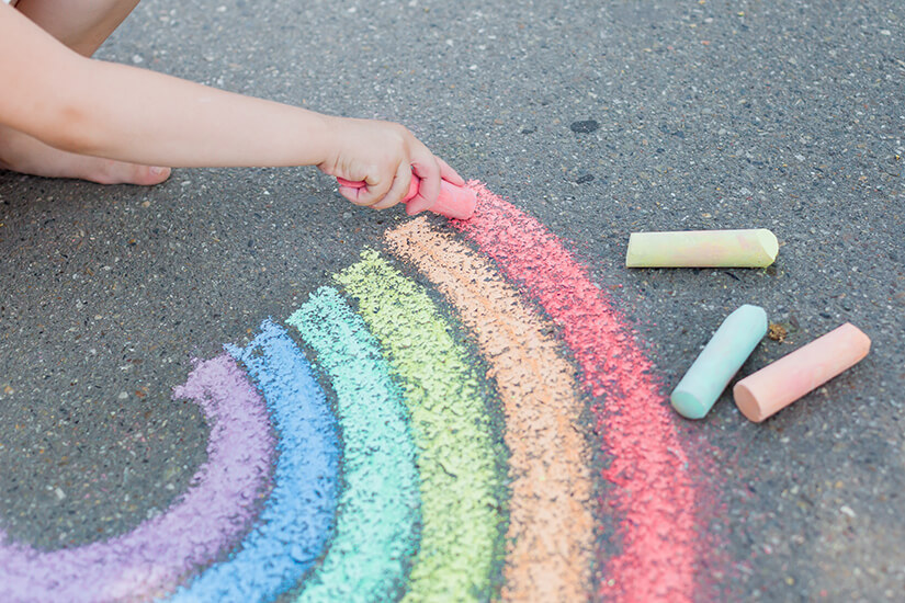 giving-back-to-the-society-HG Child drawing a colorful rainbow on asphalt with sidewalk chalk, surrounded by pastel-colored chalk pieces.