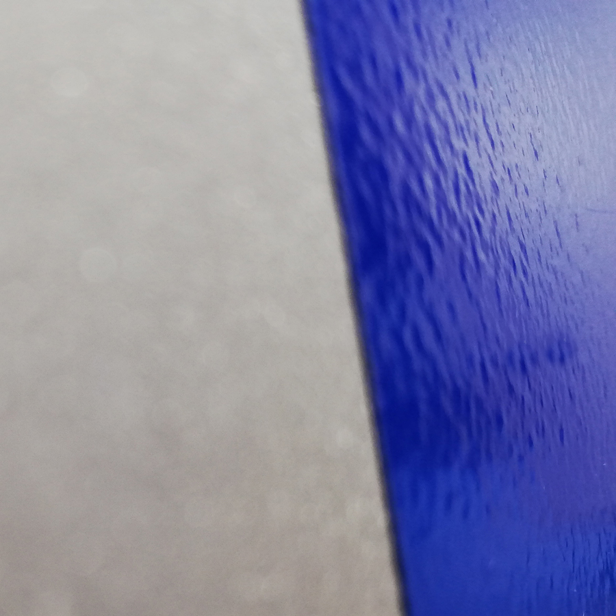 liquid-monolayer A close-up view of a square sample plate showing two distinct surface areas: one side reveals the untreated, raw metal texture, while the other side is coated with a structured, blue monolayer finish, illustrating the contrast between uncoated and coated surfaces.