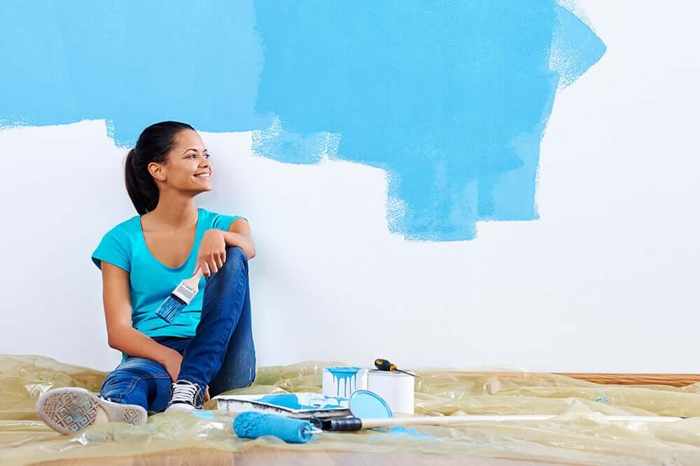wallpaint-blue Smiling woman painting interior wall light blue with roller and brush – DIY home renovation with water-based wall paint