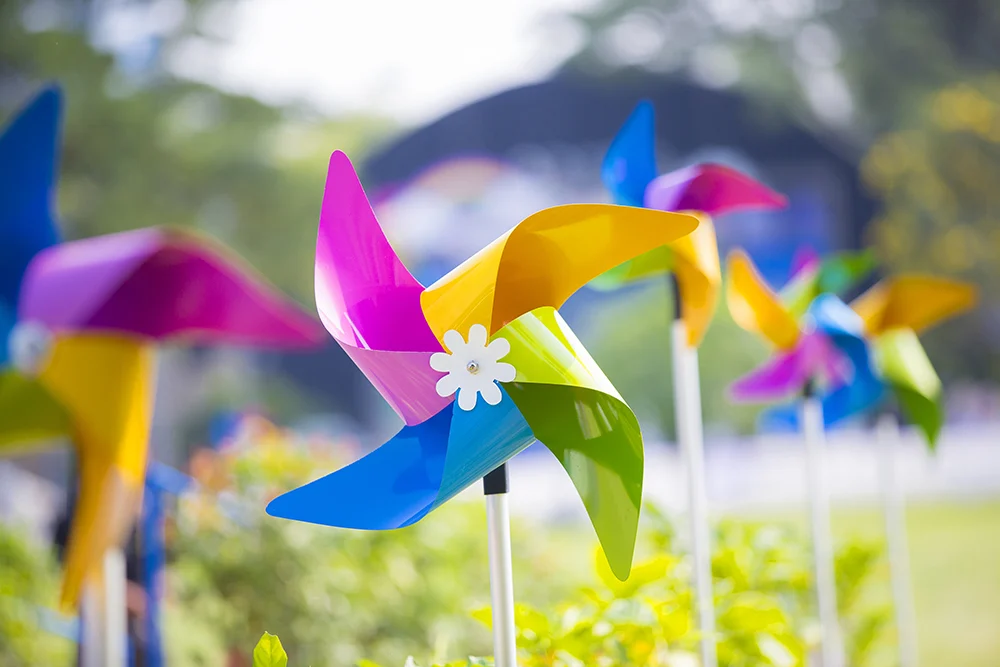 wind-sustainability-vetrnica Colorful plastic pinwheels spinning in the wind on a sunny day, set in a garden with greenery and a blurred background.